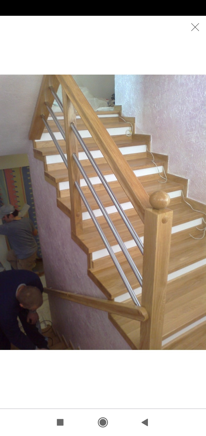 Montaż drewnianych schodów z metalowymi balustradami; widoczni dwaj pracownicy podczas instalacji, jeden przy balustradzie, drugi w tle, jasne drewno, białe podstopnice, faktura ściany.