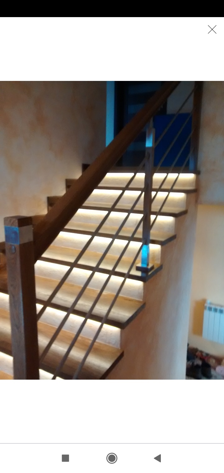 Kątowe ujęcie drewnianych schodów z podświetleniem LED na każdym stopniu i metalową balustradą, w tle ściana o fakturze piasku.