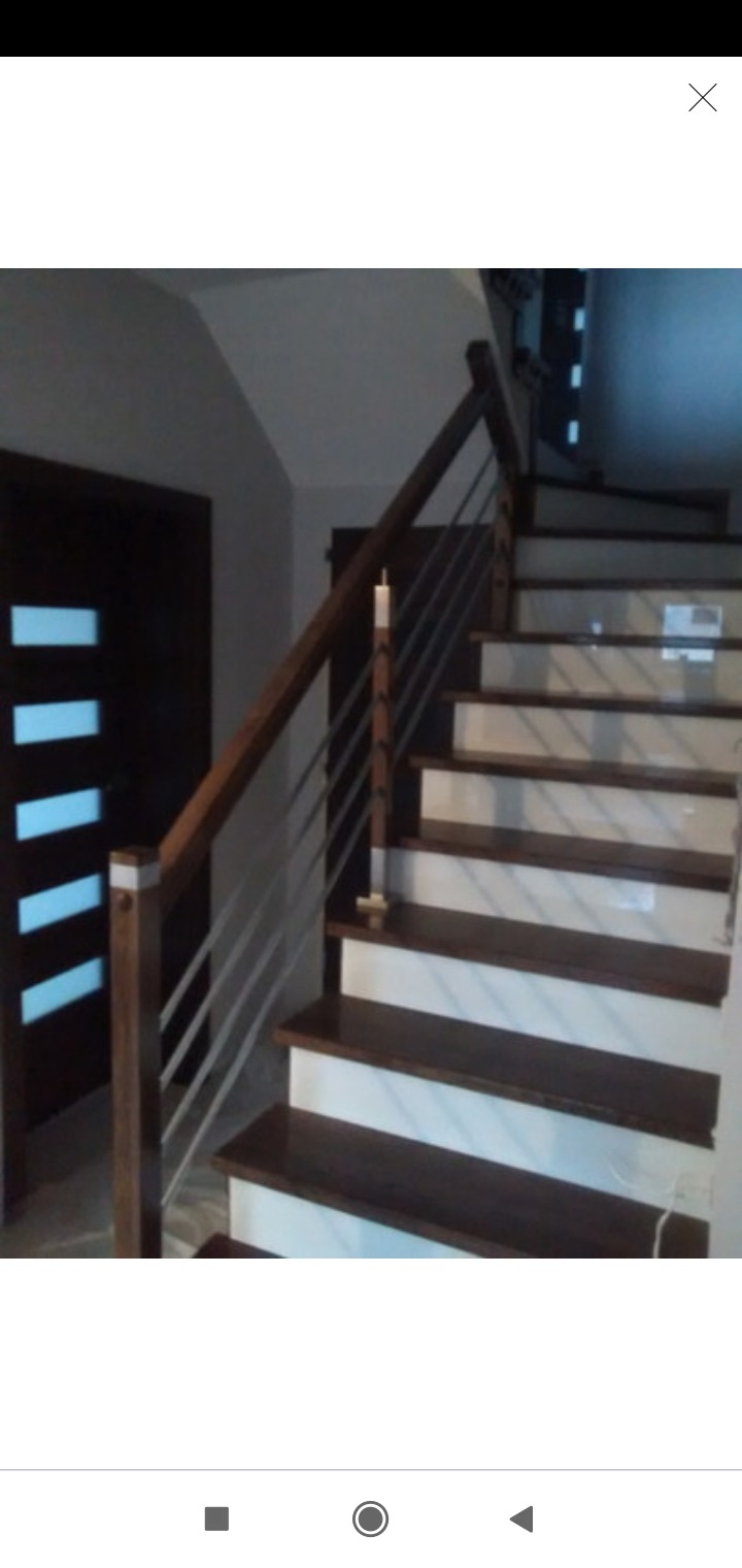 Ciemnobrązowe schody z białymi podstopnicami i metalową balustradą, widoczne z dołu, obok ciemnych drzwi z poziomymi, jasnymi szybami.