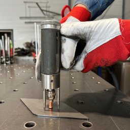 Mosqito Metal Design - Zbliżenie na proces zgrzewania punktowego blachy, widoczne elektrody urządzenia i rękawice ochronne operatora na perforowanym stole spawalniczym.