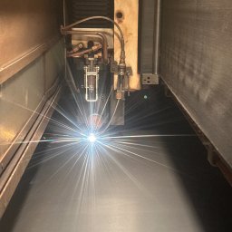 Mosqito Metal Design - Zbliżenie na proces cięcia laserowego metalu wewnątrz maszyny, z widoczną głowicą i intensywną wiązką światła.