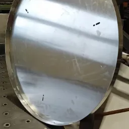 Okrągły, spawany element ze stali nierdzewnej, leżący na stole spawalniczym z otworami, widoczne ślady po spawaniu punktowym.