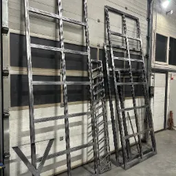 Surowe, metalowe ramy okienne z podziałami, elementy konstrukcyjne schodów oraz podstawa stołu w kształcie litery X, przygotowane do dalszej obróbki lub montażu, ustawione w hali produkcyjnej.