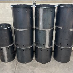 Mosqito - Ciemne, metalowe cylindry z widocznymi spoinami i ozdobnymi wycięciami w kształcie płomieni, ułożone w stosy na betonowej posadzce.