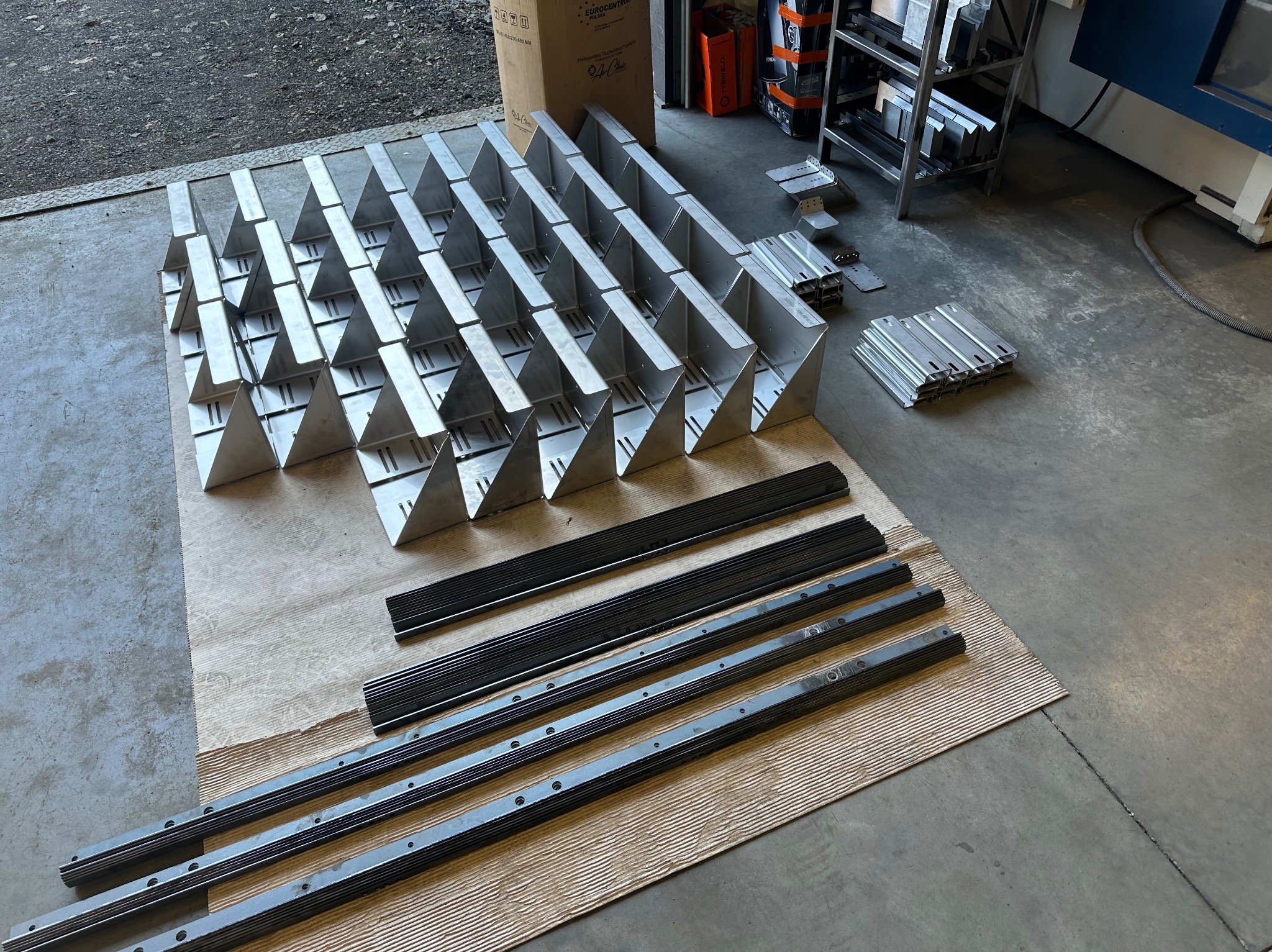 Elementy konstrukcyjne ze stali nierdzewnej, w tym kątowniki i profile z otworami, ułożone na papierze pakowym i podłodze warsztatu, przygotowane do montażu.
