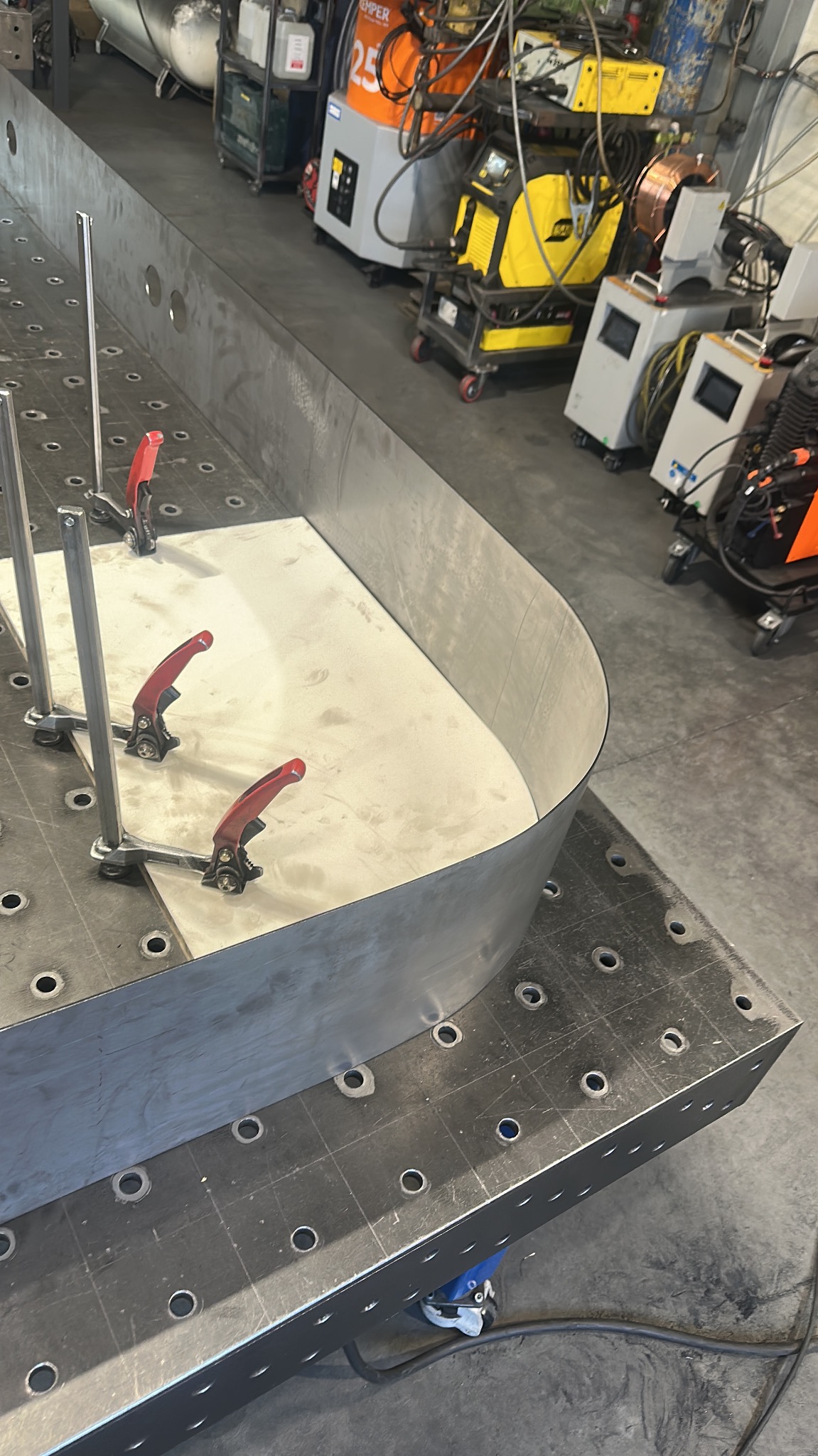 Blacha zagięta w łuk mocowana klamrami na perforowanym stole spawalniczym, w tle widoczne urządzenia spawalnicze.