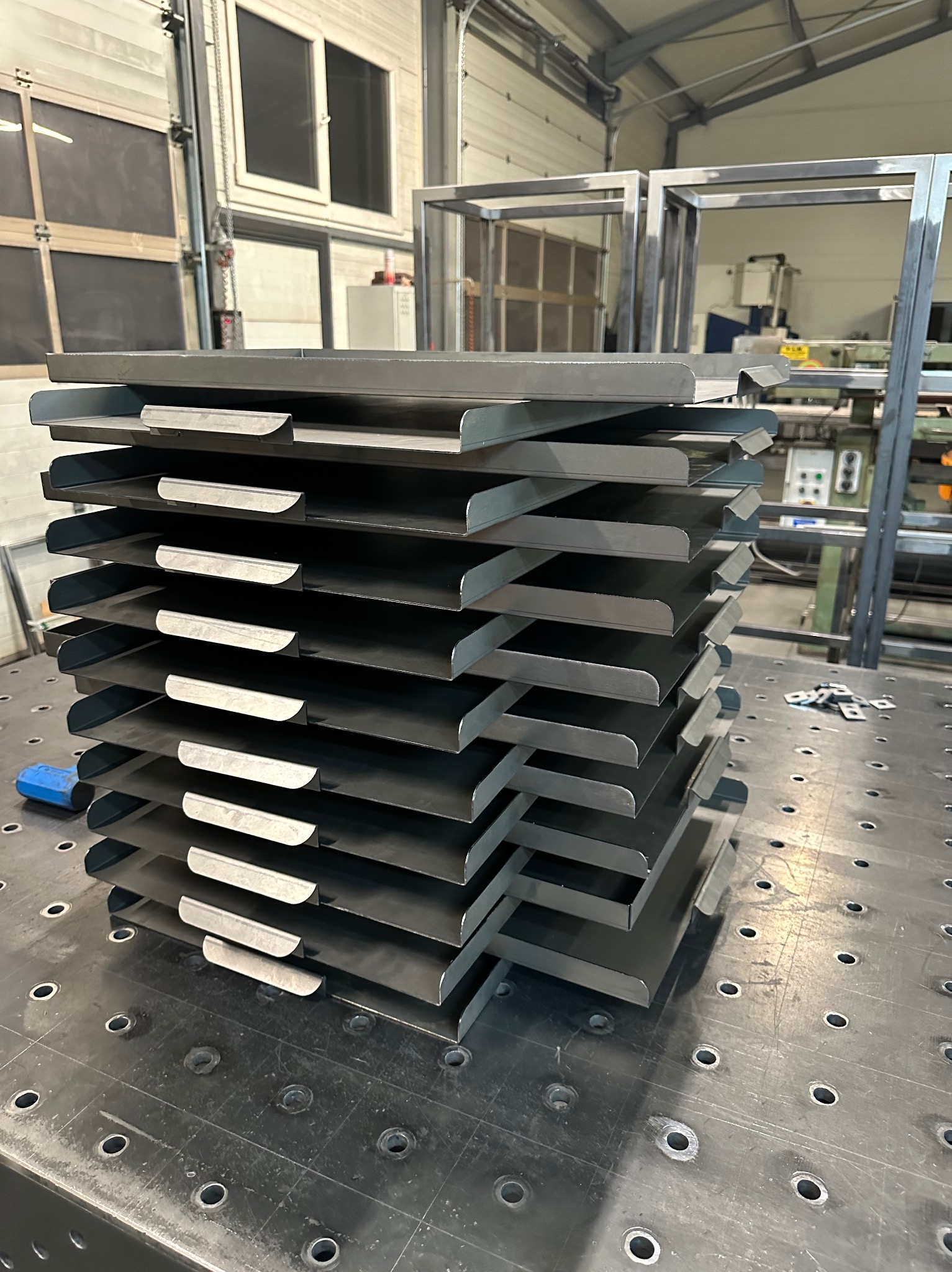 Stos precyzyjnie wyciętych i giętych metalowych elementów na perforowanym stole warsztatowym, w tle widoczne metalowe ramy i maszyny.