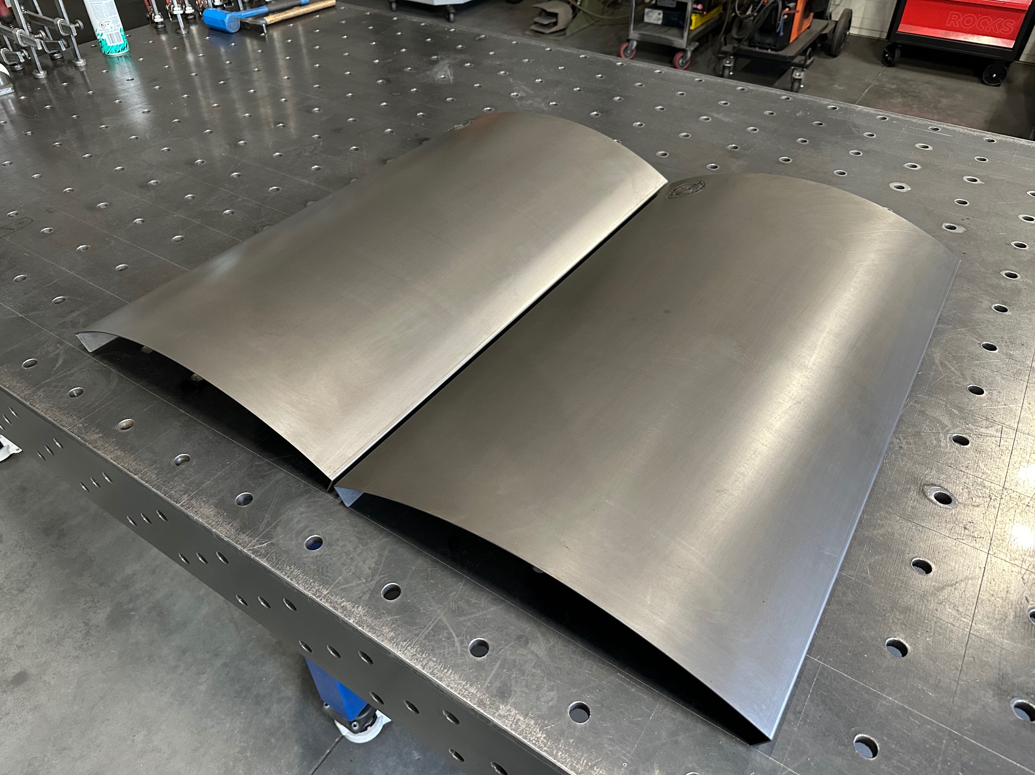 Dwie wygięte, surowe blachy z nierdzewnej stali leżące na perforowanym stole spawalniczym; widoczne narzędzia i wózki warsztatowe w tle.