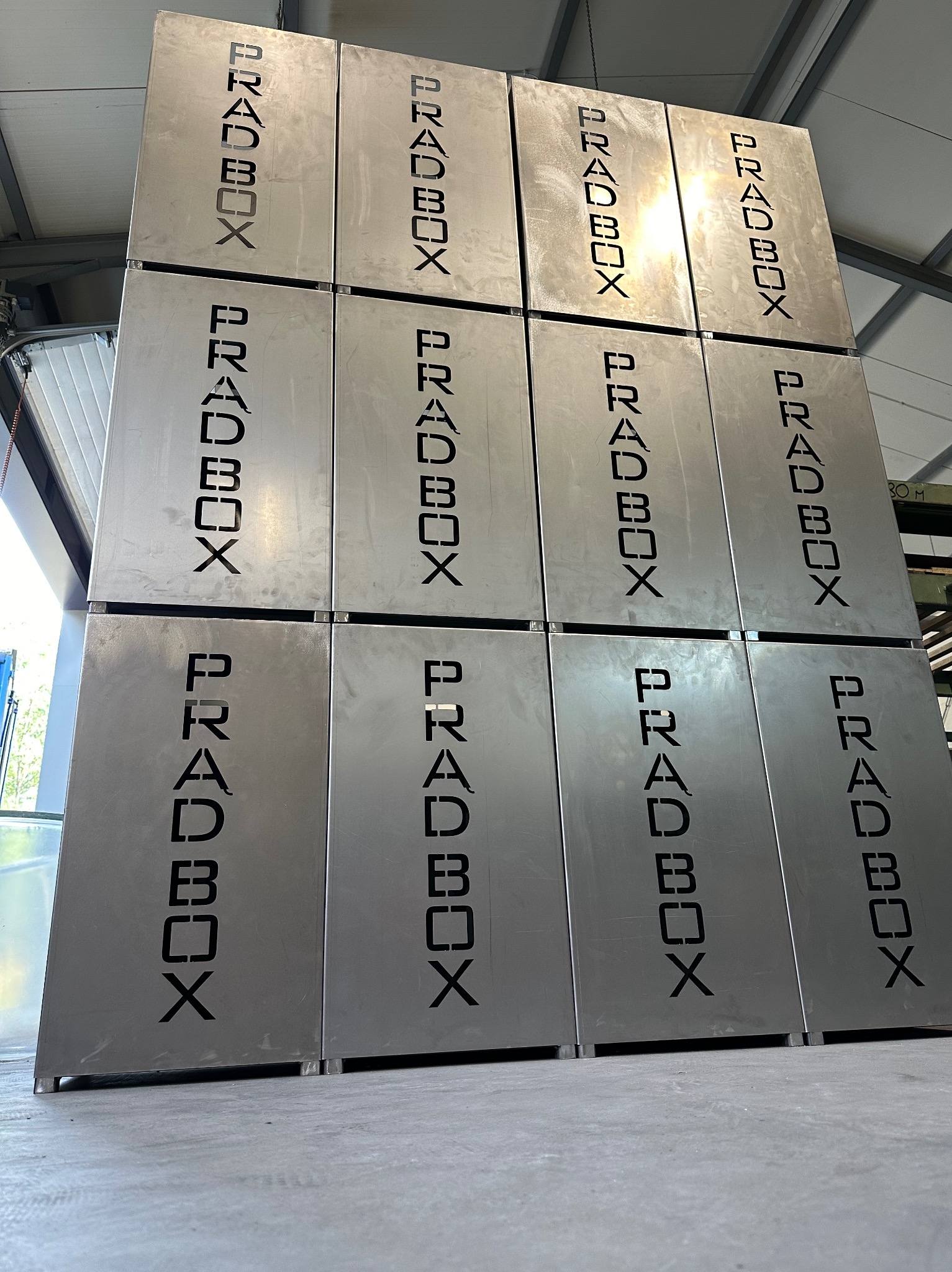 Wysoki stos identycznych, srebrnych, metalowych obudów z wyciętym laserowo napisem 'PRADBOX'. Obudowy ustawione jedna na drugiej w magazynie lub hali produkcyjnej. Widoczne złączenia między obudowami.
