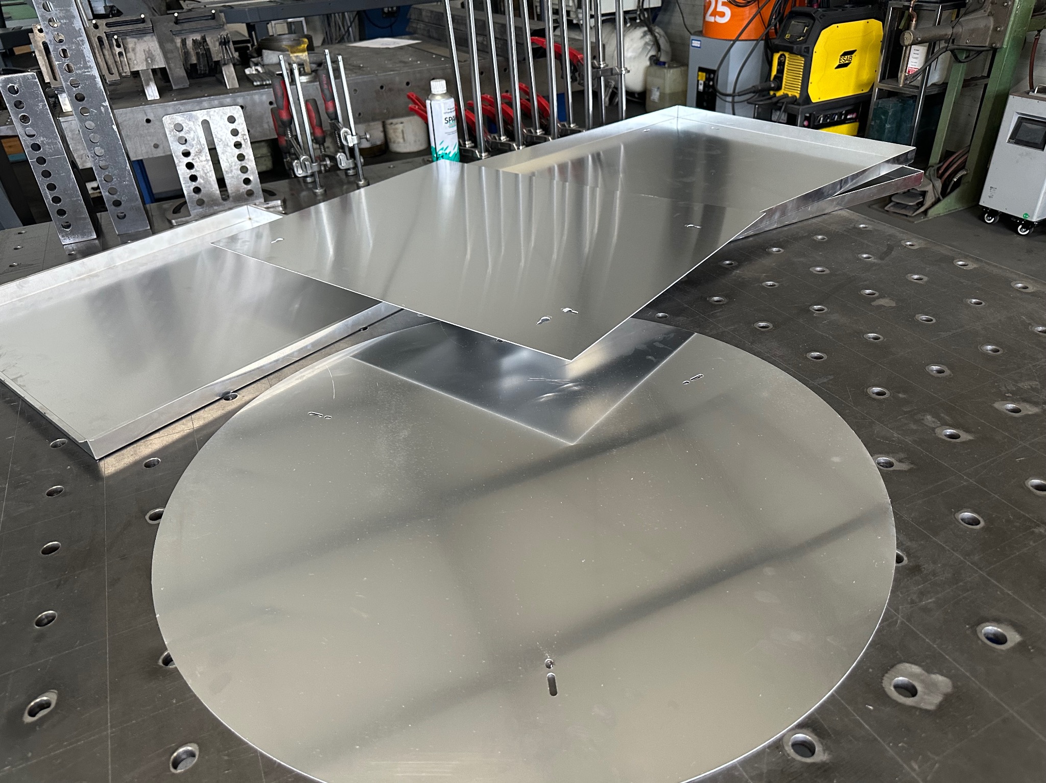 Przygotowane do dalszej obróbki, płaskie elementy metalowe (okrągły i prostokątne) z otworami, leżące na perforowanym stole warsztatowym. W tle widoczny sprzęt spawalniczy.