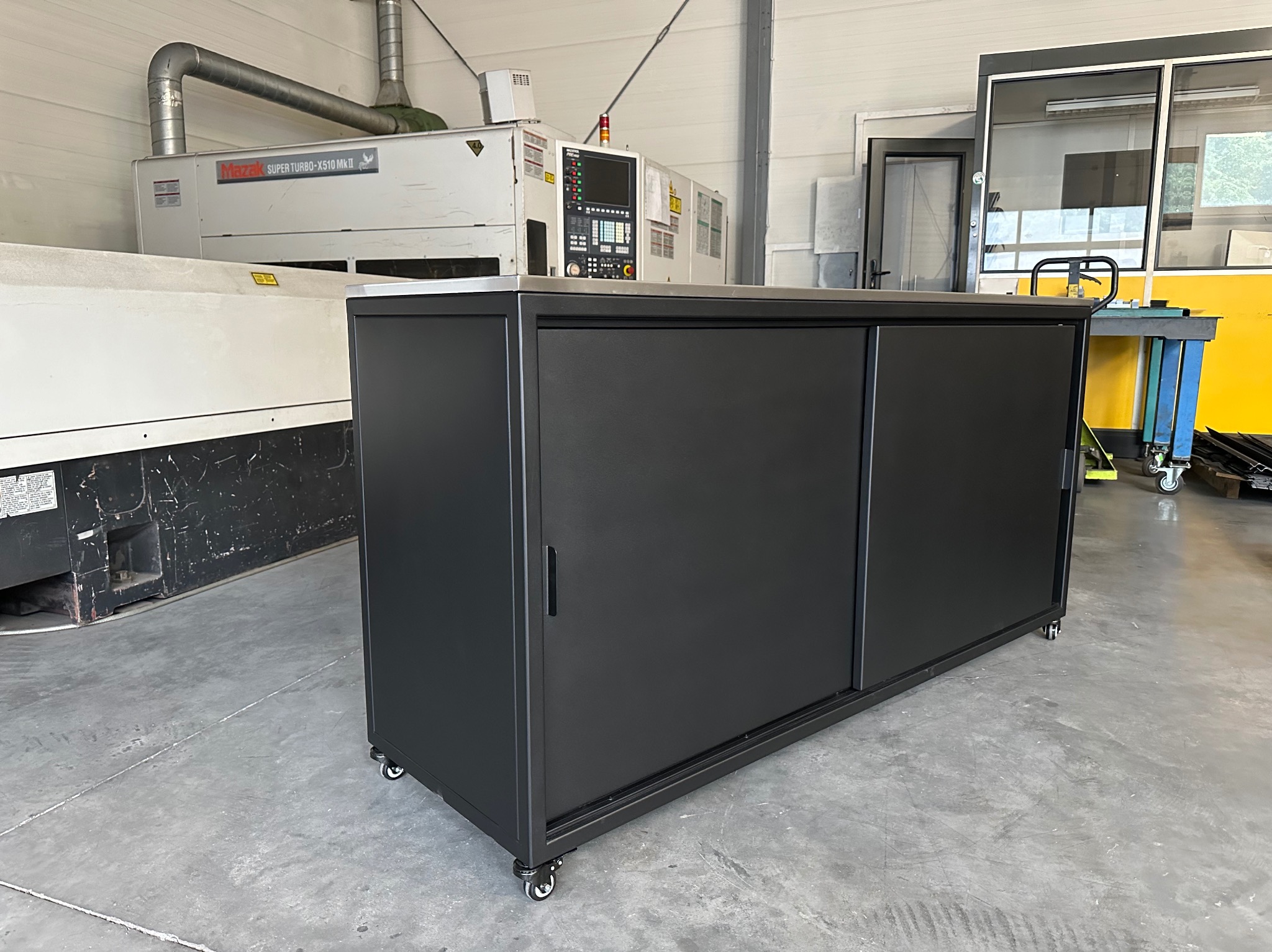 Czarna szafka warsztatowa na kółkach z przesuwnymi drzwiami, widoczna na tle maszyny CNC Mazak Super Turbo-X510 MKII w hali produkcyjnej.