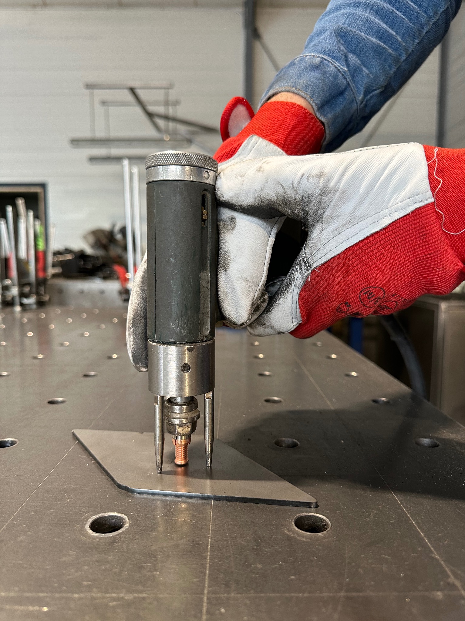 Zbliżenie na proces zgrzewania punktowego blachy, widoczne elektrody urządzenia i rękawice ochronne operatora na perforowanym stole spawalniczym.