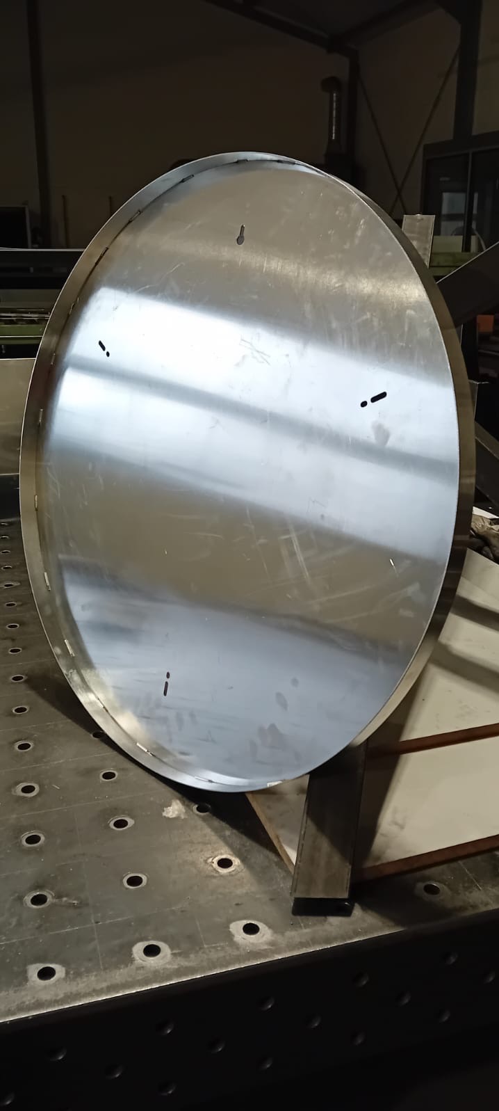 Okrągły, spawany element ze stali nierdzewnej, leżący na stole spawalniczym z otworami, widoczne ślady po spawaniu punktowym.