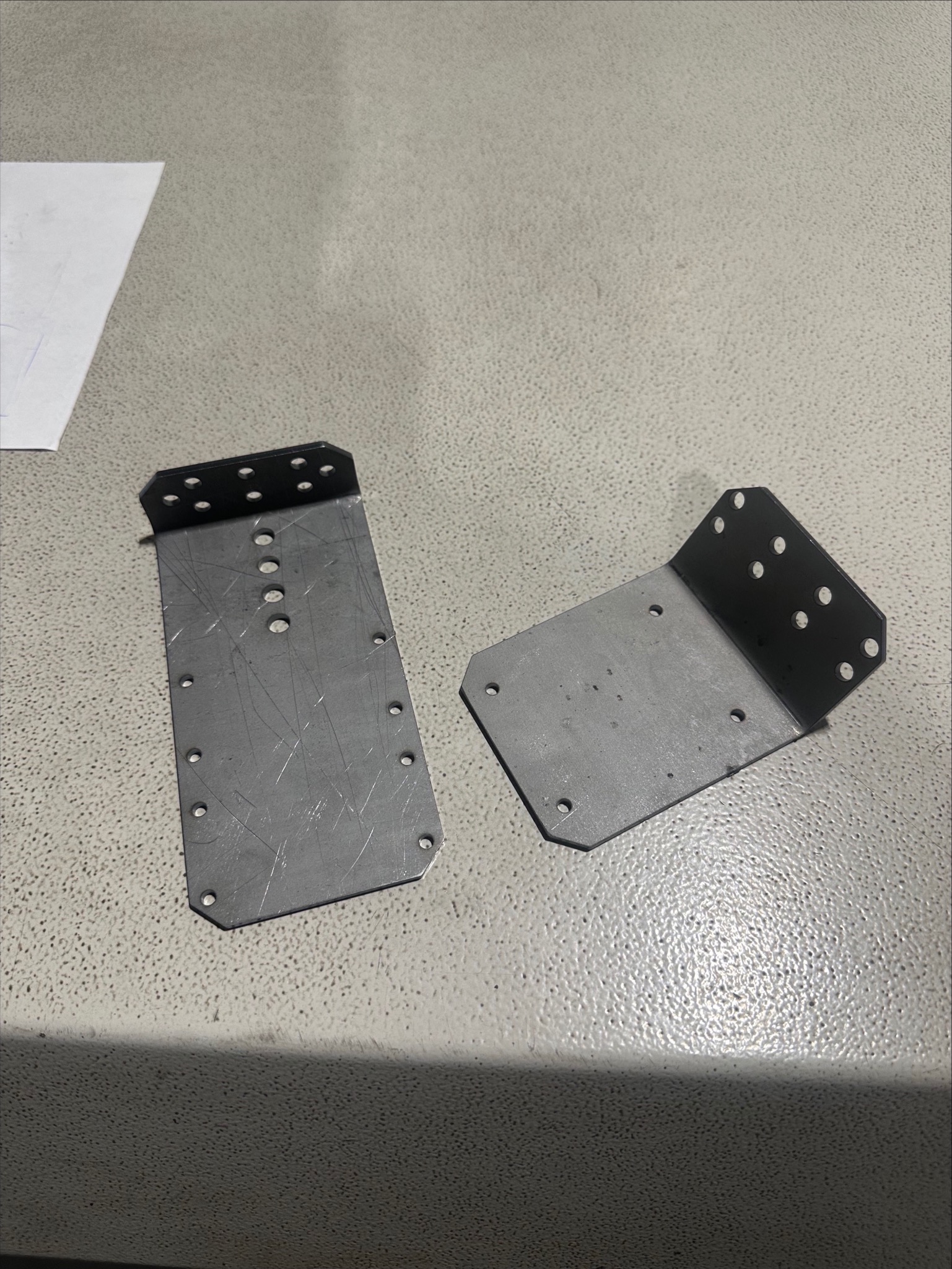 Dwa metalowe elementy z otworami, prawdopodobnie łączniki lub wsporniki, leżące na szarej powierzchni. Jeden element jest płaski z zagiętą krawędzią, drugi jest zagięty pod kątem prostym.