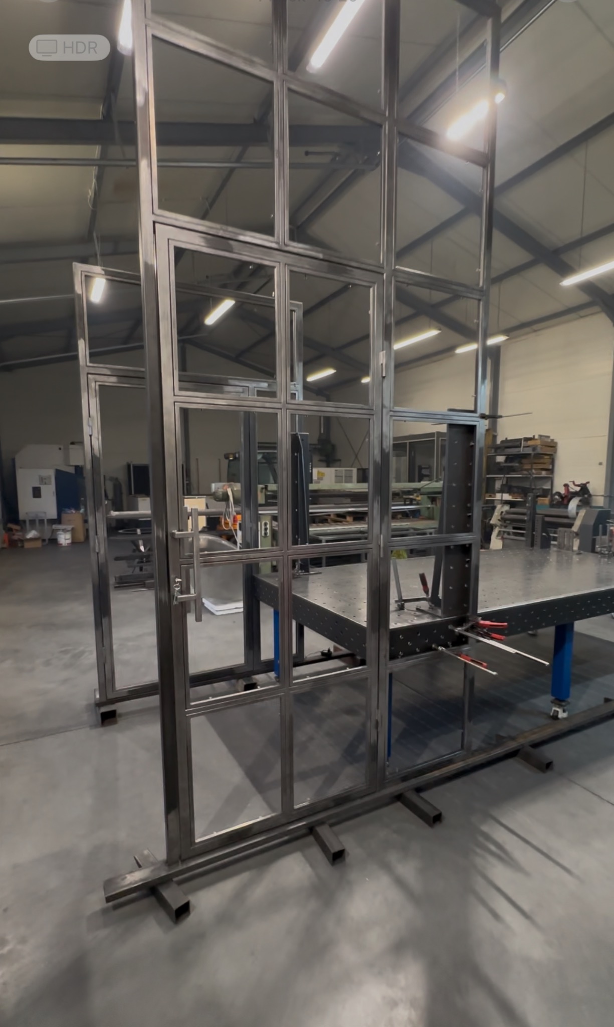 Konstrukcja metalowa z profili, prawdopodobnie ścianka działowa lub element zabudowy, w trakcie montażu w hali produkcyjnej. Widoczne spawane łączenia i elementy mocujące.