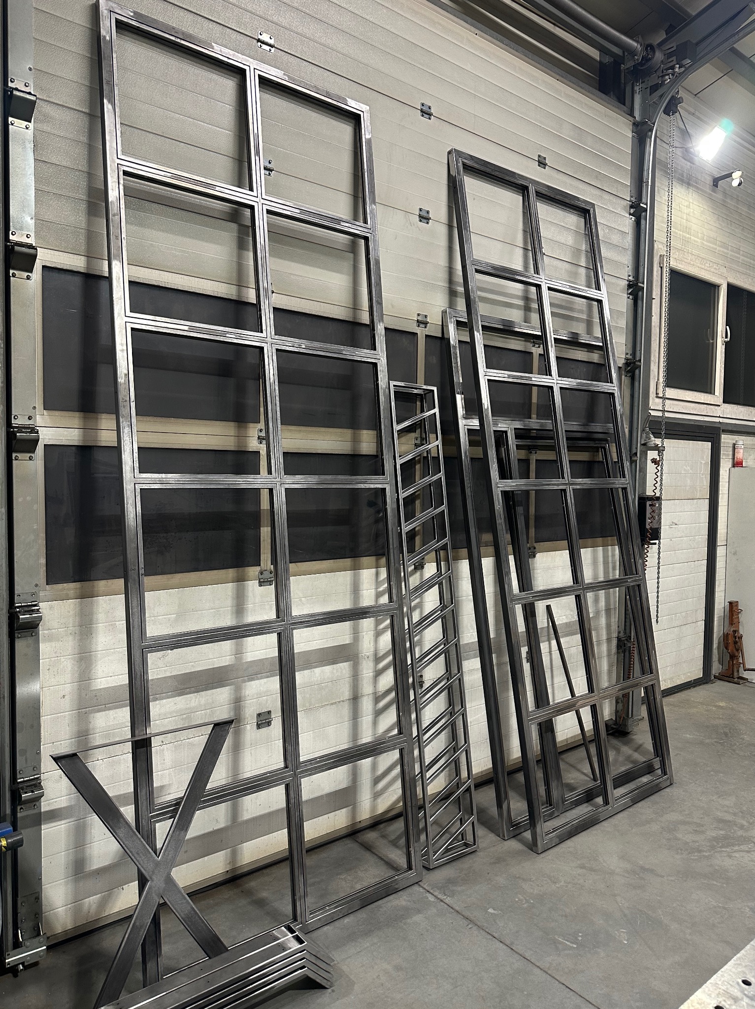 Surowe, metalowe ramy okienne z podziałami, elementy konstrukcyjne schodów oraz podstawa stołu w kształcie litery X, przygotowane do dalszej obróbki lub montażu, ustawione w hali produkcyjnej.