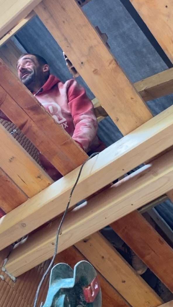 Mężczyzna w bluzie z kapturem pracuje nad drewnianą konstrukcją dachu, widok z dołu, surowy stan, widoczne deski i elementy mocujące.