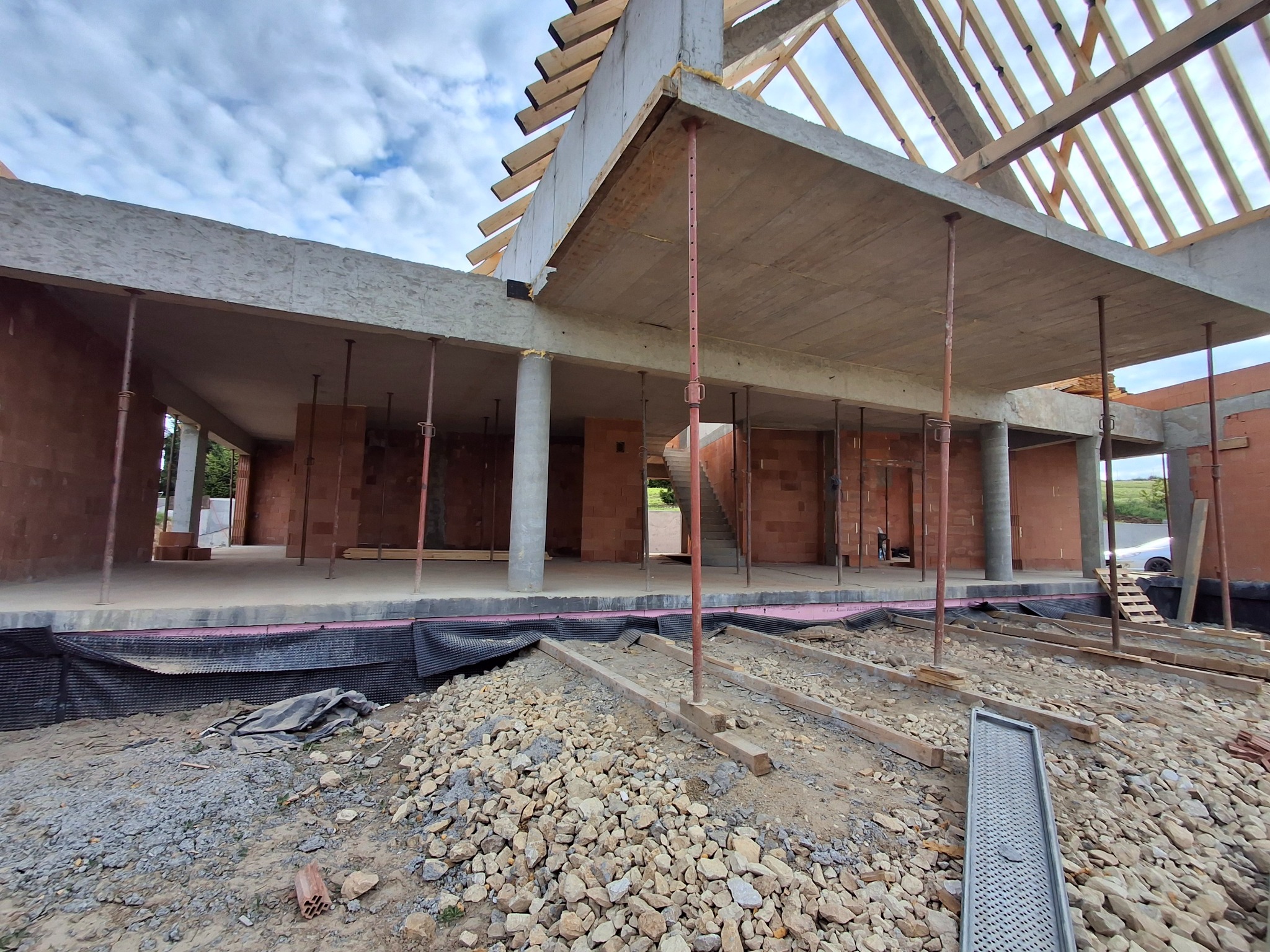 Surowy stan budynku żelbetowego w trakcie budowy, widoczne podpory stropu, ściany z czerwonej cegły, szalunki i żwir na gruncie. Perspektywa z dołu.