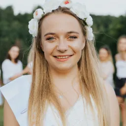 Uśmiechnięta blondynka w białej sukience z koronkowym wykończeniem, wiankiem z białych kwiatów na głowie i szarfą z napisem 'Bride to be', na tle zieleni i rozmytych postaci.