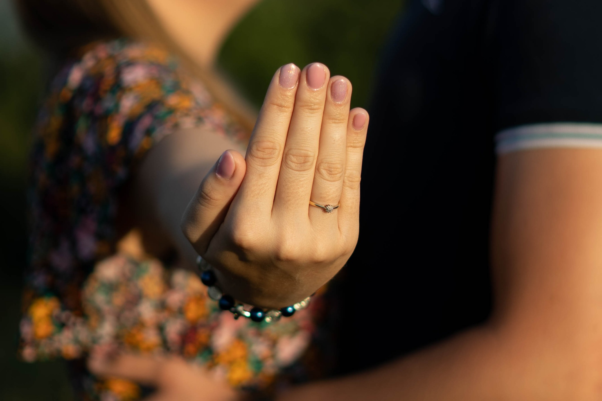 Dłoń kobiety z delikatnym pierścionkiem zaręczynowym na palcu serdecznym, bransoletka z ciemnych koralików, w tle zamazana sylwetka mężczyzny i kwiecista sukienka.