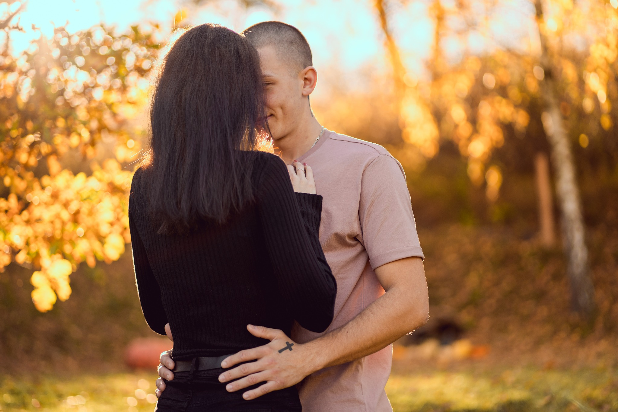 Para obejmująca się na tle jesiennego krajobrazu, mężczyzna z tatuażem krzyża na dłoni, ciepłe światło słoneczne.