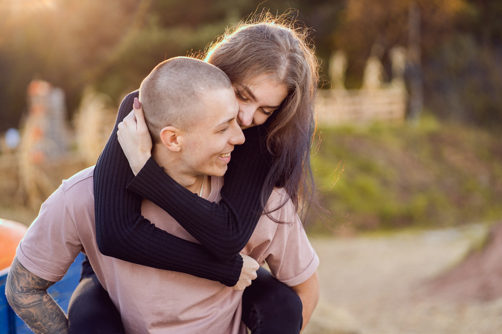 Uśmiechnięty mężczyzna z tatuażami na ramieniu nosi na plecach młodą kobietę, która obejmuje go za szyję w promieniach zachodzącego słońca.