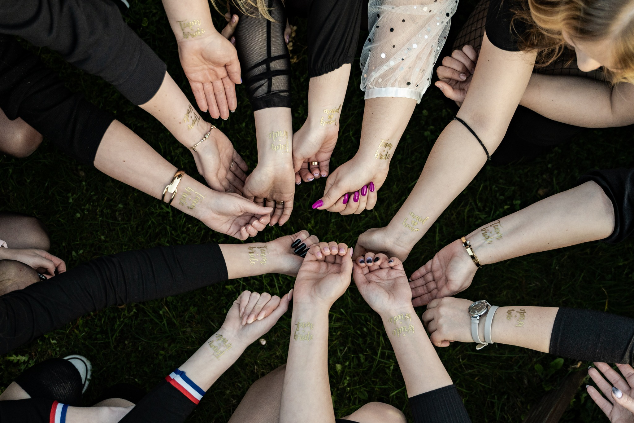 Grupa kobiet trzymająca się za ręce, z tymczasowymi złotymi tatuażami z napisami 'Team Bride' i 'Maid of Honor' na przedramionach, na tle zielonej trawy.