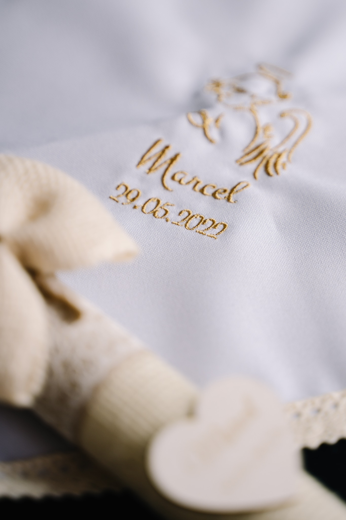 Biała szatka do chrztu z haftowanym imieniem Marcel, datą 29.05.2022 i motywem anioła w kolorze złotym, obok leży kremowa świeca z serduszkiem.