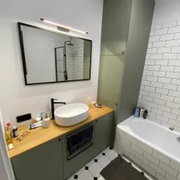 Nowoczesna łazienka z szafkami w odcieniach oliwkowej zieleni, drewnianym blatem, owalną umywalką i lustrem w czarnej ramie; w tle białe płytki i kabina prysznicowa z czarnymi profilami.