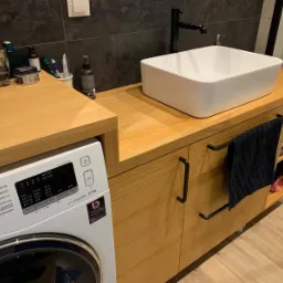 Łazienka z drewnianym blatem, na którym stoi biała, kwadratowa umywalka i czarna bateria; obok pralka zabudowana pod blatem; drewniane szafki z czarnymi uchwytami i półkami na ręczniki.