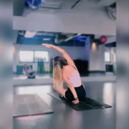 Kobieta w sportowym stroju wykonuje skłon boczny na macie do ćwiczeń w przestronnym, jasnym pomieszczeniu z widocznymi instalacjami na suficie.