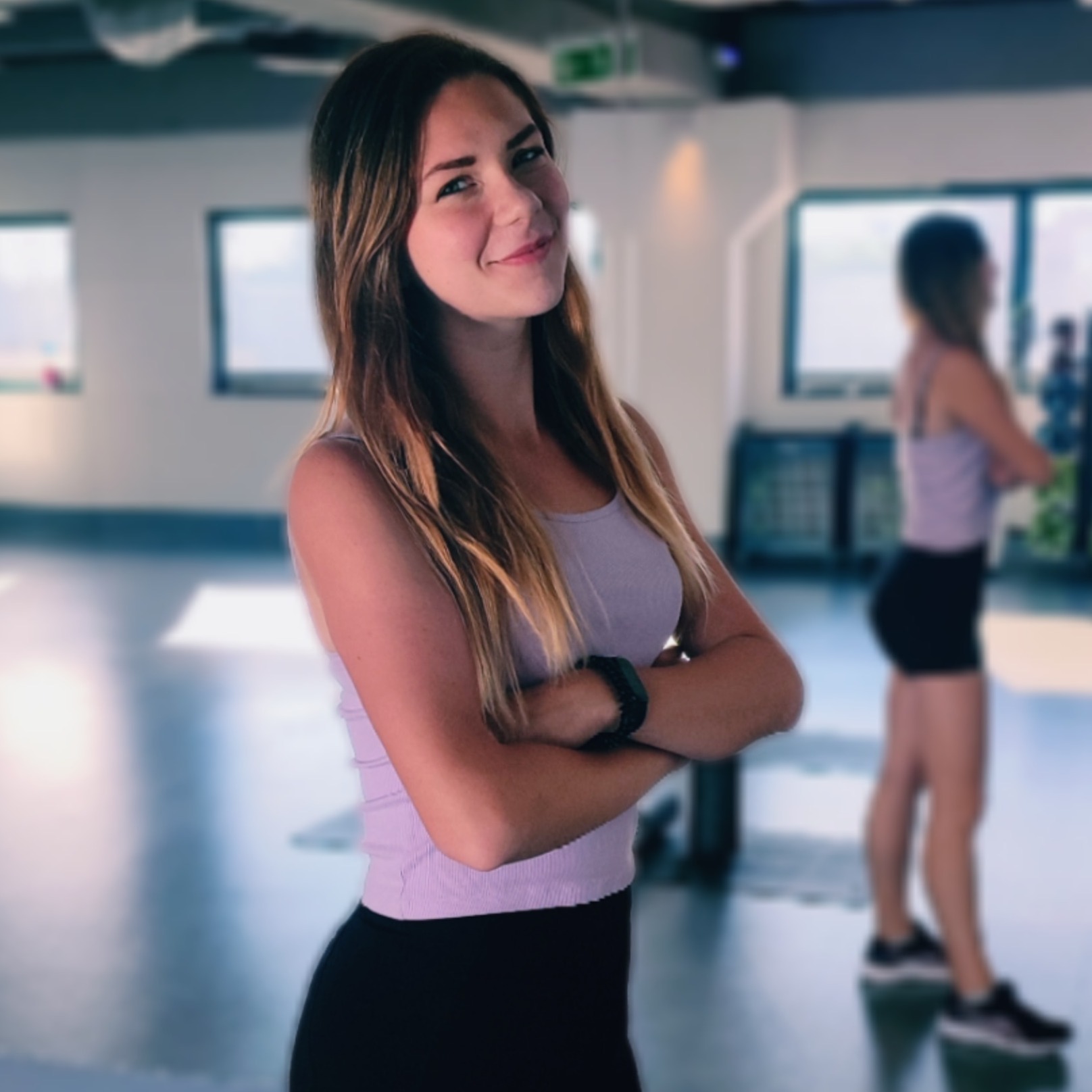 Uśmiechnięta młoda kobieta z blond włosami, ubrana w fioletowy top i czarne legginsy, stoi z założonymi rękami na tle jasnego, minimalistycznego wnętrza sali do ćwiczeń, gdzie w tle widać drugą osobę.