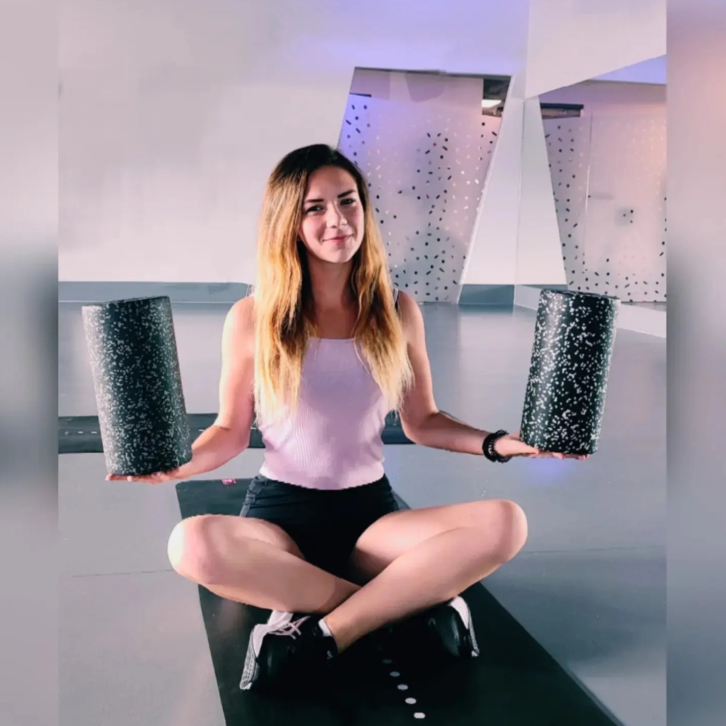 Kobieta siedząca na macie do ćwiczeń, trzymająca dwa czarne, teksturowane wałki do automasażu w dłoniach, uśmiechająca się do kamery.
