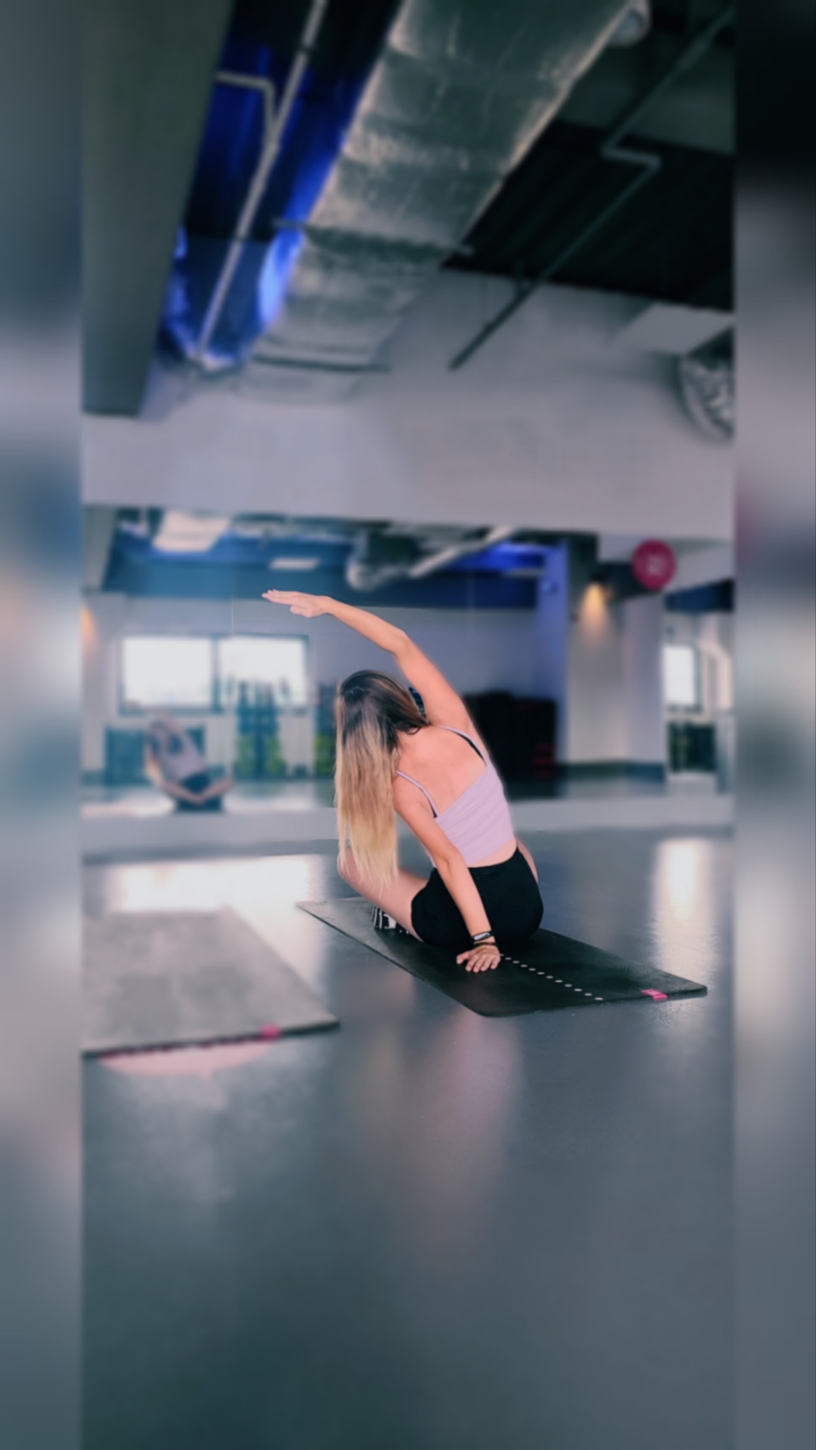 Kobieta w sportowym stroju wykonuje skłon boczny na macie do ćwiczeń w przestronnym, jasnym pomieszczeniu z widocznymi instalacjami na suficie.