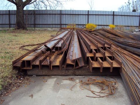 Stos dwuteowników HEB 160 i wiązki prętów zbrojeniowych składowane na zewnątrz, widoczne ślady korozji, w tle ogrodzenie z blachy falistej i drzewo.