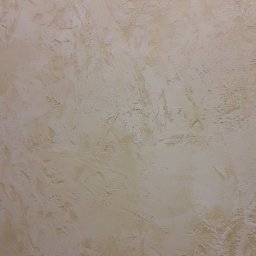 A.L. Andrii Chylin - Beżowa ściana z wyraźną, nieregularną fakturą, imitująca naturalny kamień lub tynk strukturalny, oświetlona z góry, ukazująca subtelne cieniowanie i detale powierzchni.