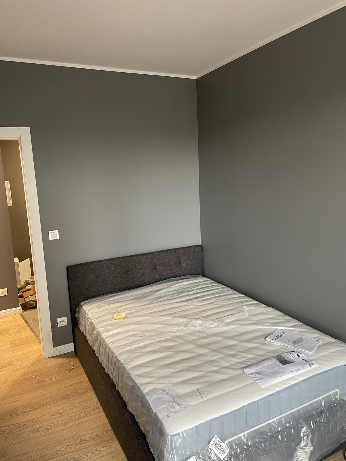 Świeżo pomalowane szare ściany w sypialni, z nowym łóżkiem w folii i instrukcjami na materacu, widoczne gniazdka elektryczne.
