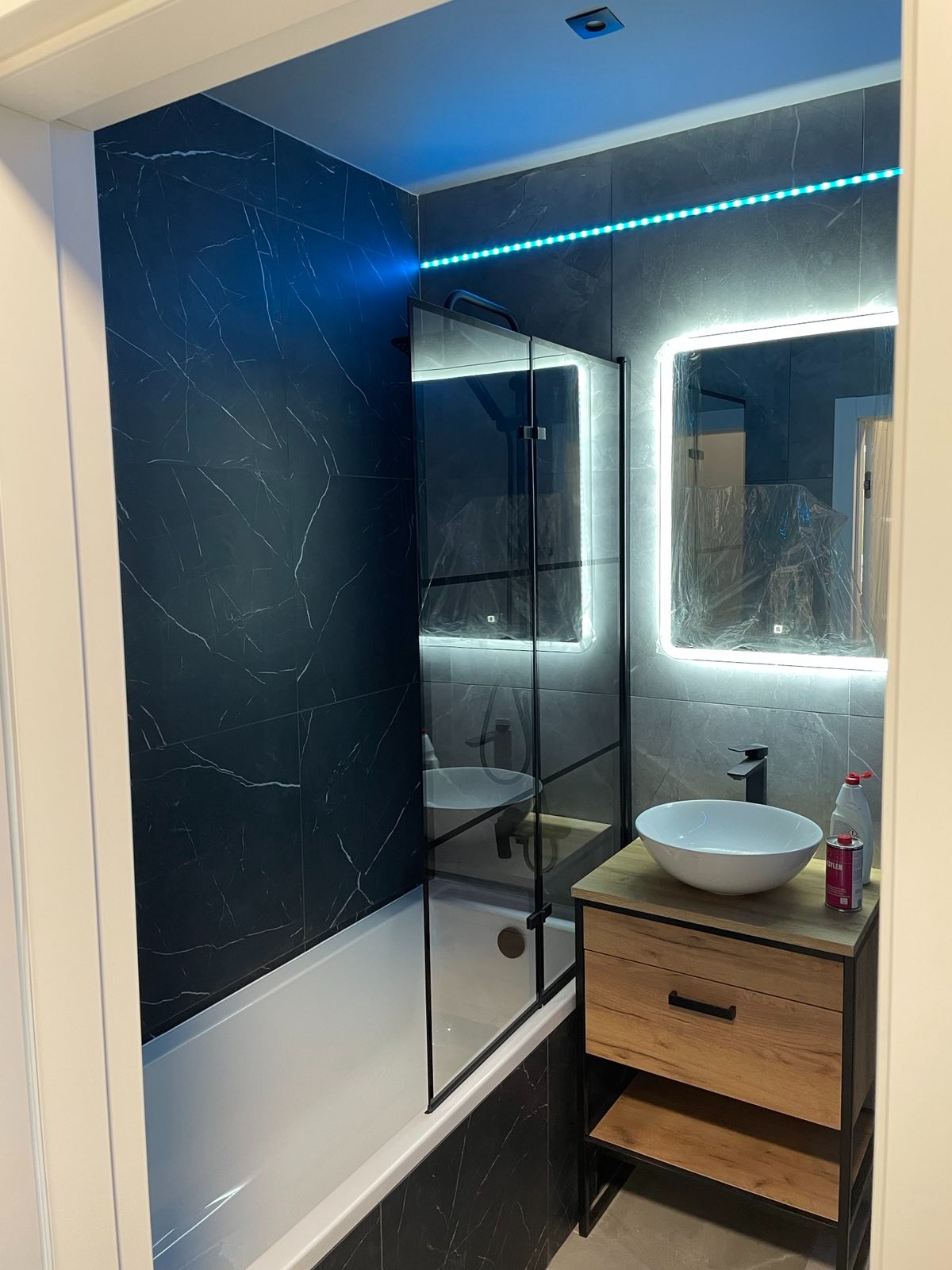 Nowoczesna łazienka z czarnymi płytkami imitującymi marmur, oświetleniem LED, białą wanną z parawanem, umywalką nablatową na szafce z drewnianym frontem i lustrem z podświetleniem LED.