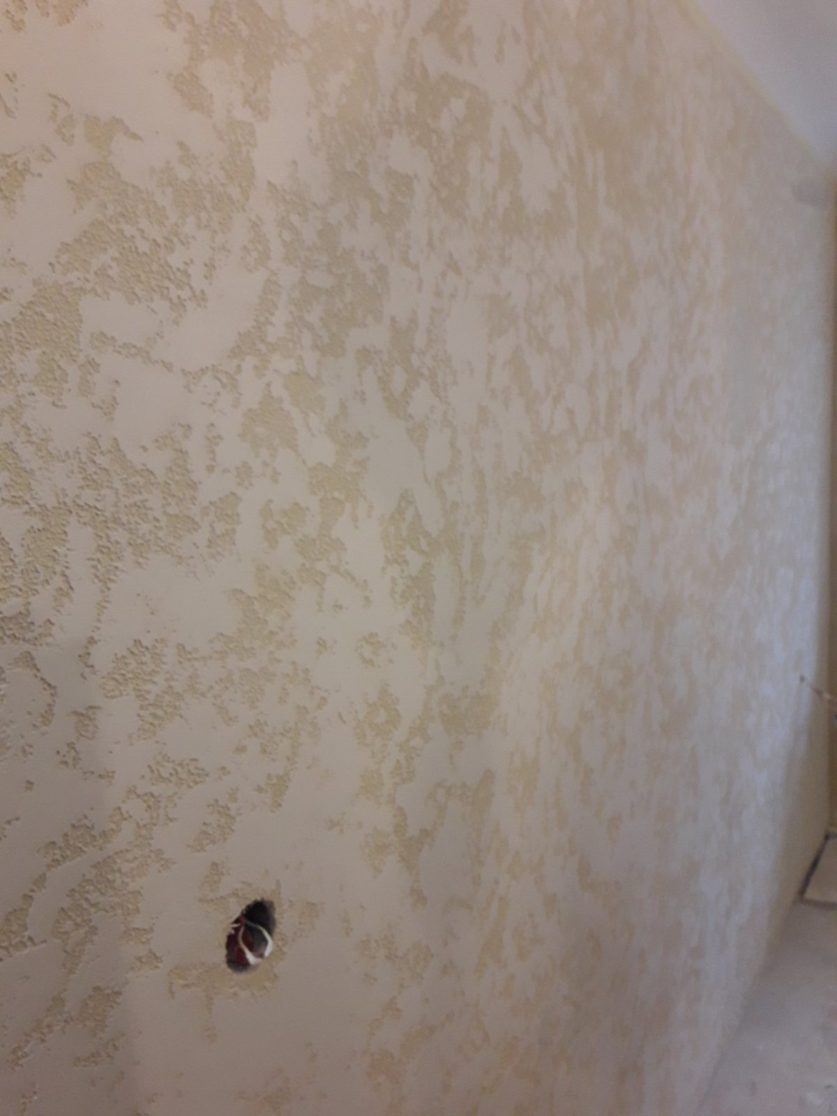 Beżowa ściana z dekoracyjną strukturą, widoczny otwór na gniazdko elektryczne z przewodami.