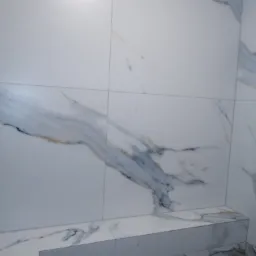 Ściana i półka wyłożone białymi płytkami imitującymi marmur z szarymi i złotymi żyłami, widoczne fugi.