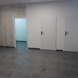Wnętrze w trakcie wykańczania: szare płytki podłogowe, dwie białe zamontowane drzwi z czarnymi klamkami, ściana częściowo wykończona gładzią, widoczne niebieskie znaczniki na podłodze.