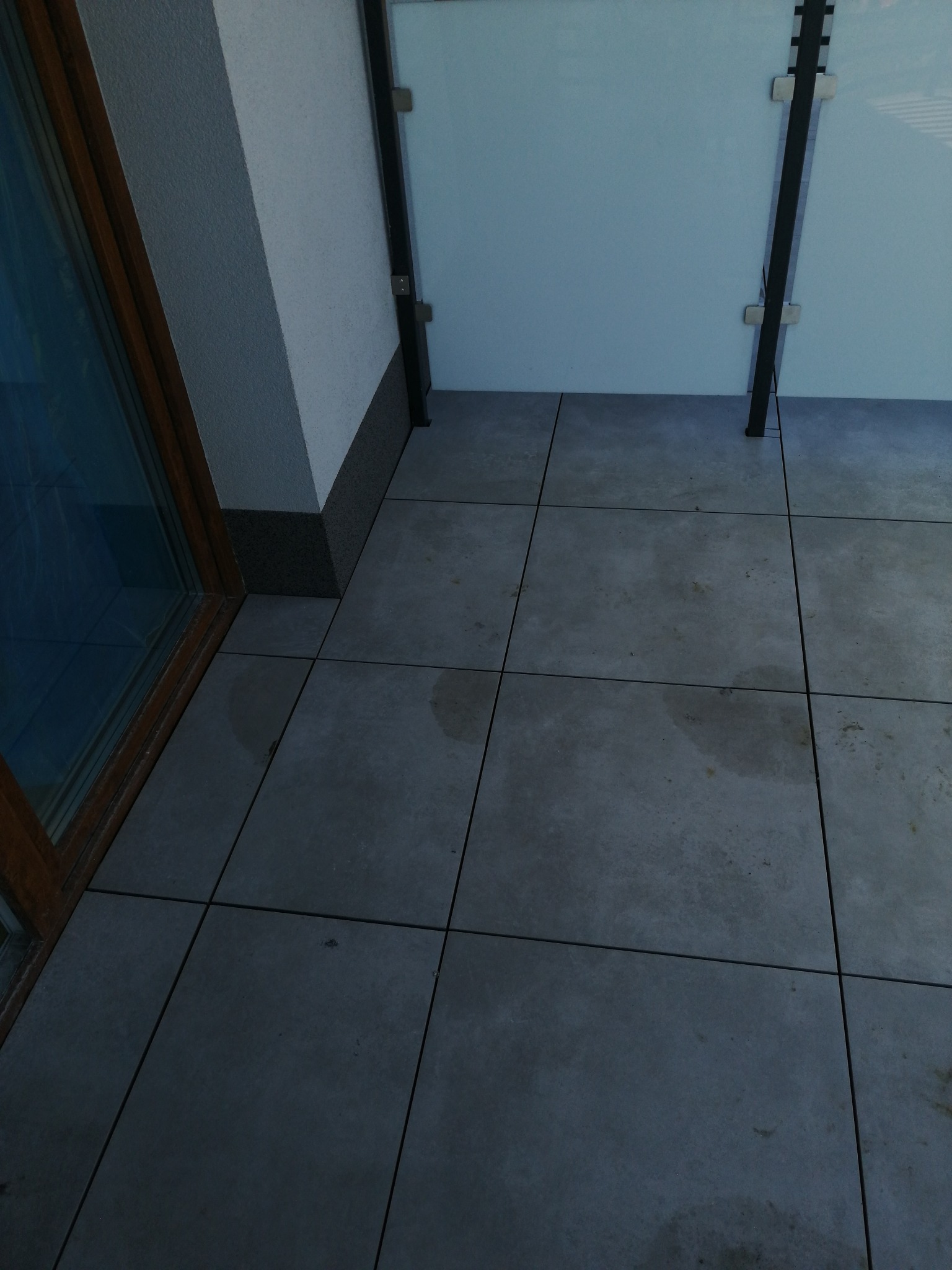 Szare, kwadratowe płytki podłogowe na balkonie z przeszkloną balustradą; widoczne ślady wilgoci na powierzchni płytek.