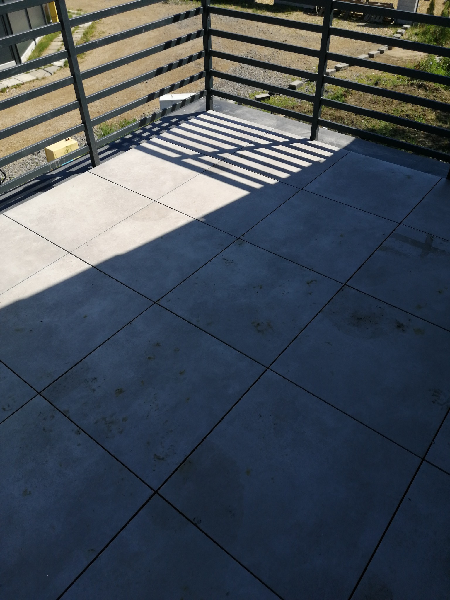 Wykończony balkon z szarymi płytkami gresowymi, metalową balustradą i widocznymi cieniami od konstrukcji zadaszenia.