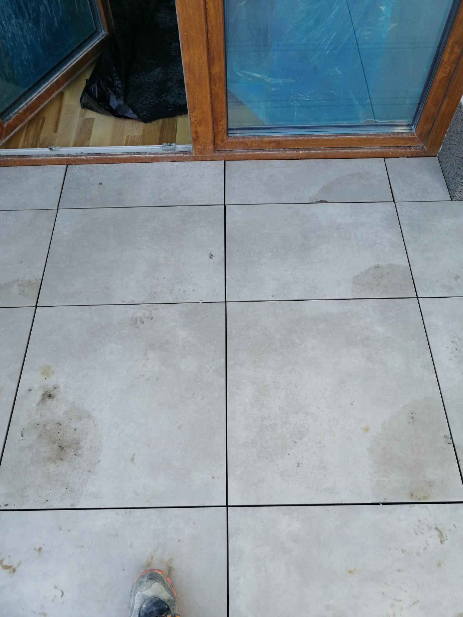 Szare płytki podłogowe na balkonie, widoczne ślady zabrudzeń, otwarte okno balkonowe z drewnianą ramą, w tle zabezpieczona folią podłoga wewnątrz pomieszczenia, fragment buta w dolnym rogu.