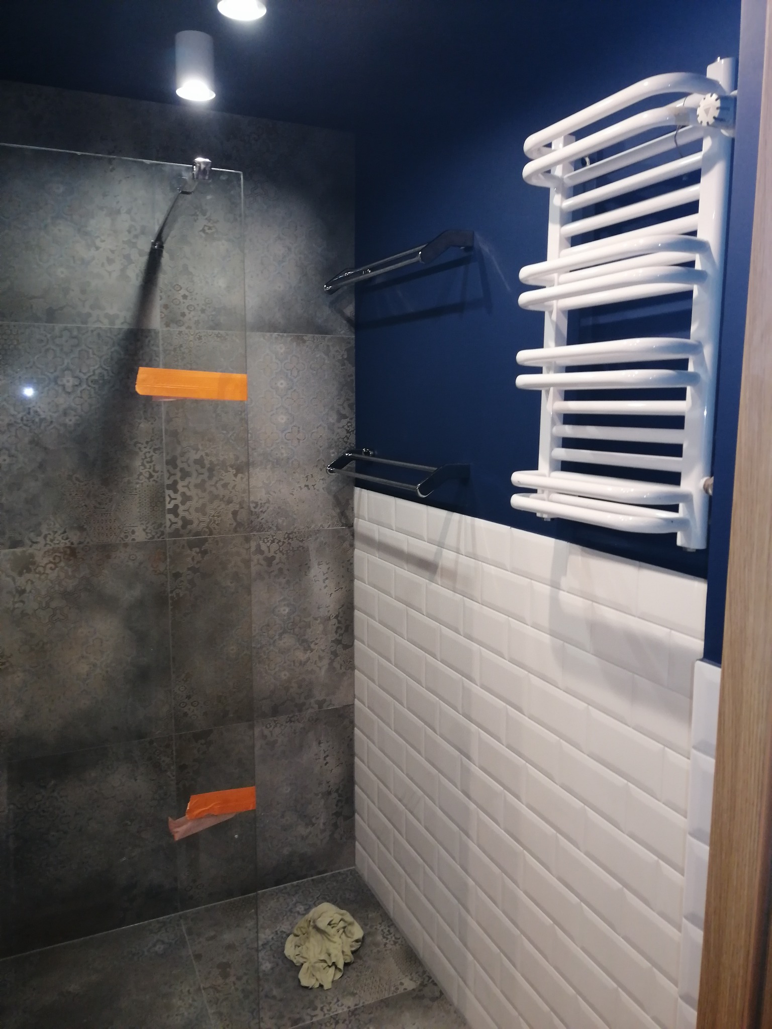 Nowoczesna łazienka z szarymi płytkami o wzorzystej fakturze, kabiną prysznicową z pomarańczowymi zabezpieczeniami, białymi płytkami typu metro i białym grzejnikiem drabinkowym na granatowej ścianie.