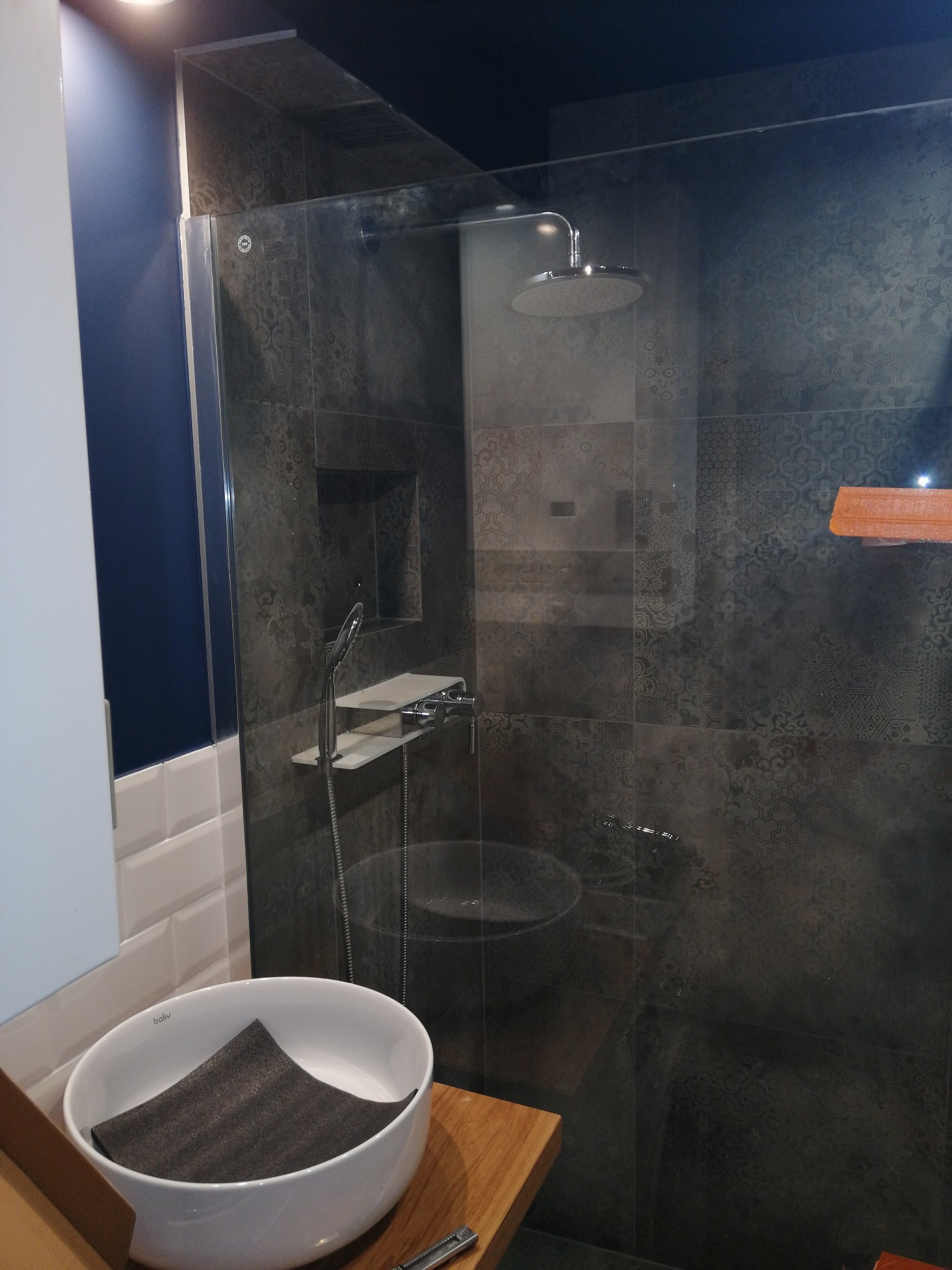 Nowoczesna łazienka z kabiną prysznicową typu walk-in, wykończona ciemnymi płytkami z dekoracyjnym wzorem, okrągłą umywalką nablatową i deszczownicą.