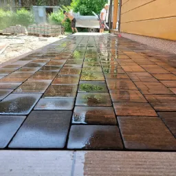 Świeżo ułożona, mokra kostka brukowa w odcieniach brązu, odbijająca niebo. W tle pracownicy i materiały budowlane, dom z drewnianą elewacją. Ujęcie z niskiej perspektywy.