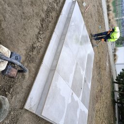 Mateo bruk - Prace brukarskie w toku: robotnik wyrównuje podłoże pod jasnoszare płyty chodnikowe. Widoczna ubijarka i narzędzia na ziemi. Praca na zewnątrz, w tle roślinność.