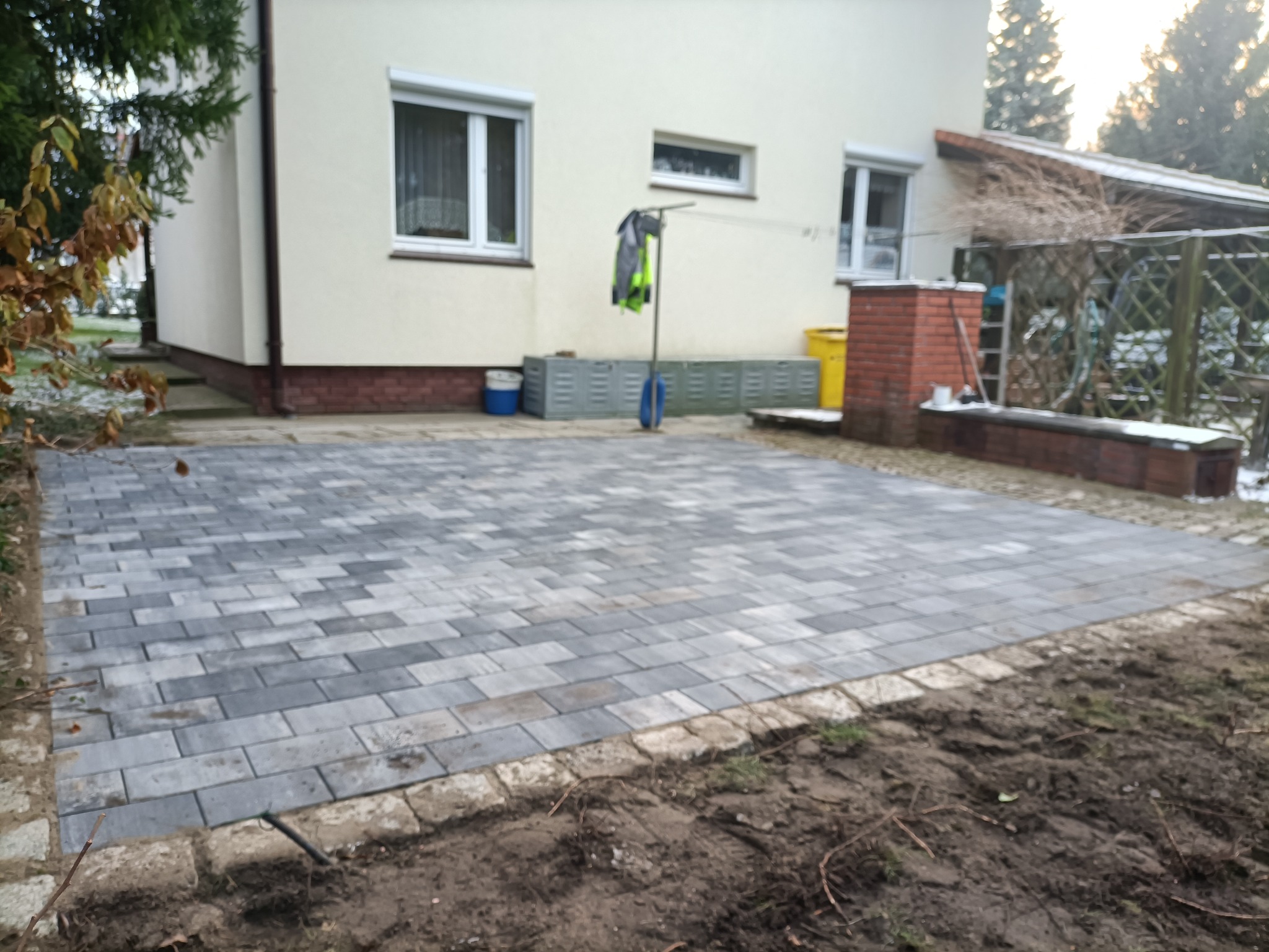 Ułożona szara kostka brukowa na podwórku domu z beżową elewacją. Widoczne obrzeża z jasnej kostki. W tle drzewa i krzewy.