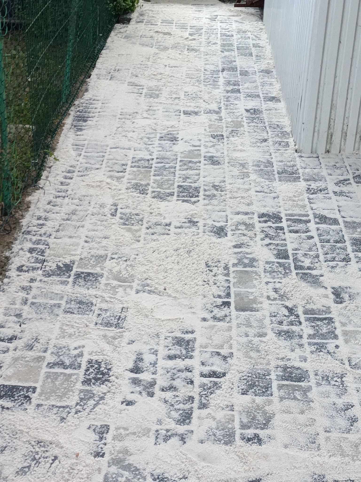 Świeżo ułożona kostka brukowa, zasypana piaskiem, widoczna z góry, obok zielona siatka ogrodzeniowa i białe deski.